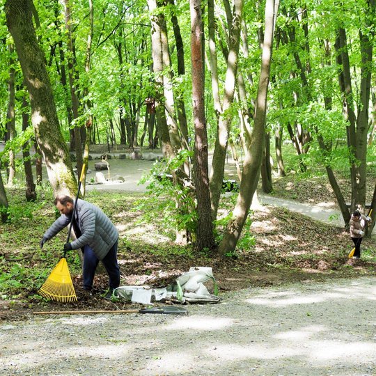 Vyras su grėbliu surenka lapus miške prie tako, šalia jo – maišas su surinktomis atliekomis. Kiek tolėliau moteris taip pat darbuojasi tarp medžių, apsupta žalumos ir saulės spindulių.