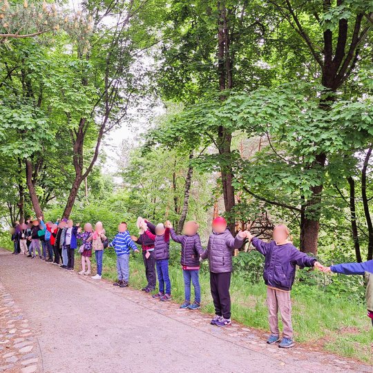 nuotraukoje – vaikai stovi išsidėstę ilga gyva grandine miško takeliu, laikosi už rankų. Tai sukuria bendrystės ir vienybės įspūdį. Nuotrauka perteikia komandinę veiklą ir bendrą patirtį.