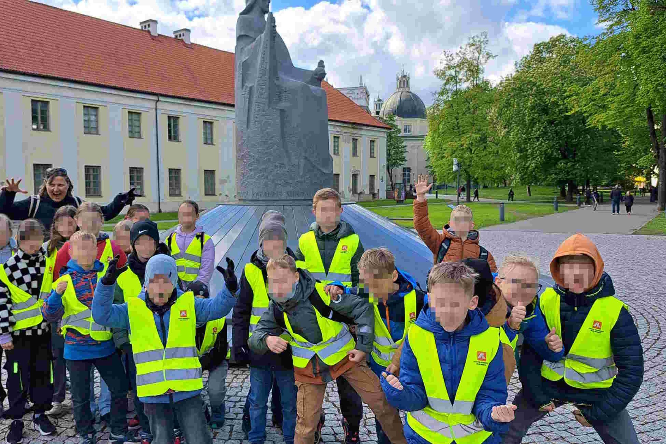 Vaikai su ryškiaspalvėmis liemenėmis pozuoja prie karaliaus Mindaugo paminklo Vilniaus pilių teritorijoje edukacijos metu.