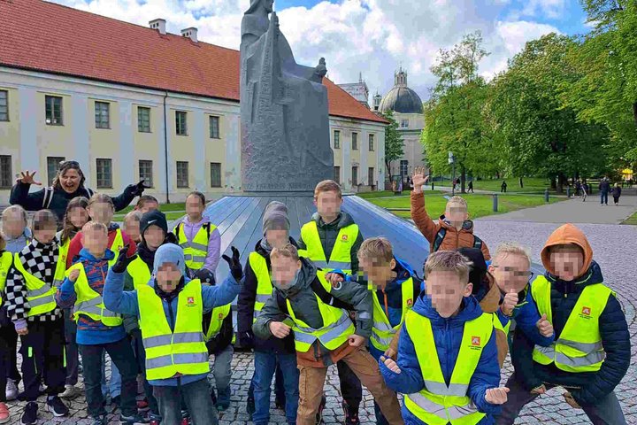 Vaikai su ryškiaspalvėmis liemenėmis pozuoja prie karaliaus Mindaugo paminklo Vilniaus pilių teritorijoje edukacijos metu.