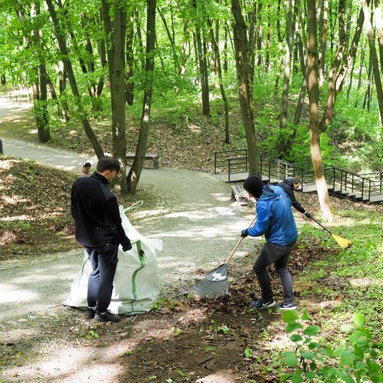 Trys žmonės valo miško pakraštį – du grėbia lapus, o vienas laiko atidarytą maišą. Fone matyti vingiuotas takas, suolelis ir nedidelis tiltelis tarp medžių.