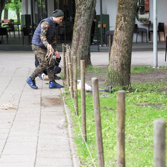 Vyras prie lauko kavinės tempia virvę tarp įkastų kuoliukų, žymėdamas ribą tarp šaligatvio ir vejos. Greta auga medžiai, o tolumoje matyti stalai ir kėdės.