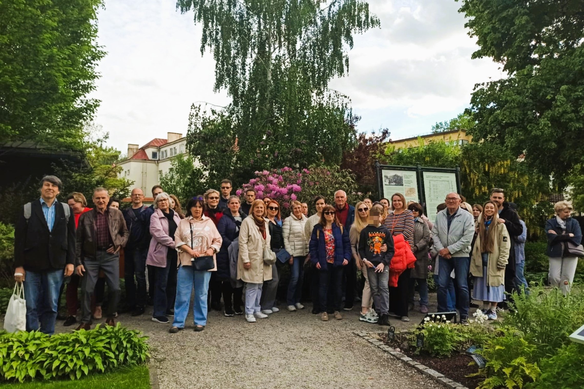 Grupė žmonių stovi žydinčiame sode prie rožinių rododendrų krūmo. Už jų matyti informaciniai stendai su tekstu ir žemėlapiu, o fone – medžiai ir keli pastatai. Žmonės žiūri į kamerą, kai kurie šypsosi. Tai ekskursijos dalyviai Vilniaus pilių teritorijoje.