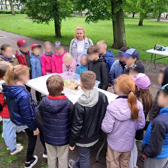 Nuotraukoje – lauko edukacinė veikla. Aplink stalą, ant kurio stovi smėlio piliakalnio modelis, susibūrę vaikai klausosi vieno iš vaikų pasakojimo. Vaikai dėvi spalvingus, šiltesnius lauko drabužius, kas rodo, jog veikla vyksta vėsesniu metų laiku. Fone m