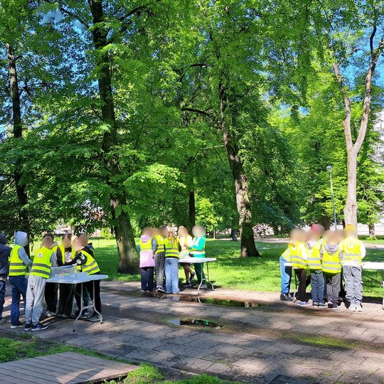 Nuotraukoje – grupė vaikų, pozuojanti prie kupolo formos stiklinės pavėsinės žalioje, medžiais apsuptoje aplinkoje. Visi vaikai dėvi ryškias geltonas saugos liemenes, o už jų matomas stendas su užrašu „Piliakalniai“ bei raudonas stovas su vėliava „VILNIAU