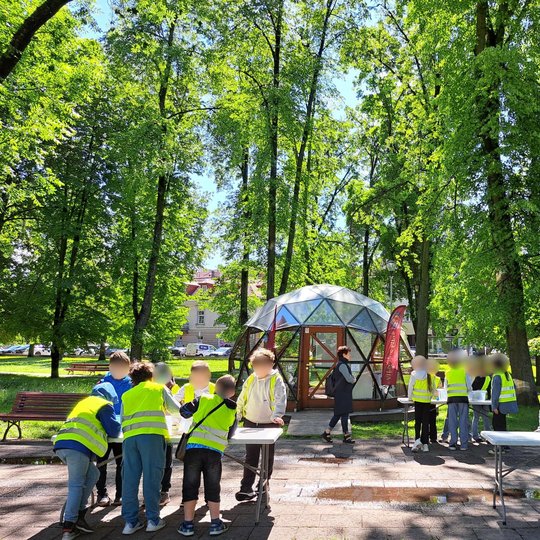 Nuotraukoje – grupė vaikų, pozuojanti prie kupolo formos stiklinės pavėsinės žalioje, medžiais apsuptoje aplinkoje. Visi vaikai dėvi ryškias geltonas saugos liemenes, o už jų matomas stendas su užrašu „Piliakalniai“ bei raudonas stovas su vėliava „VILNIAU