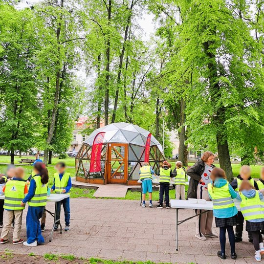 Nuotraukoje matyti grupė vaikų, vilkinčių ryškiaspalves liemenes, dalyvaujančių veiklose lauke. Vaikai susiskirstę į grupeles ir dirba prie stalų. Fone – žymus Vilniaus simbolis, Vilniaus pilių Kupolas. Aplinka žalia, su daug medžių – tai Vilniaus pilių t