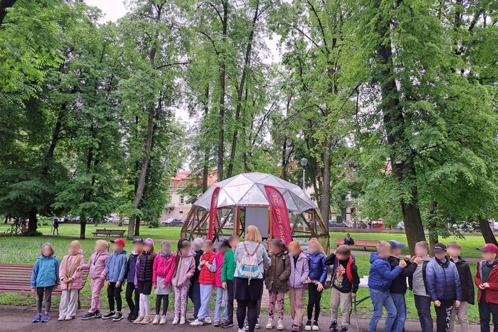 Nuotraukoje – edukacinė veikla vykstanti lauke, Vilniaus pilių teritorijoje, prie pat Gedimino pilies kalno. Vaikai sustoję vienas šalia kito, stovi prie Vilniaus pilių kupolo. Jie ruošiasi statyti piliakalnių modelius nedidelėse grupėse