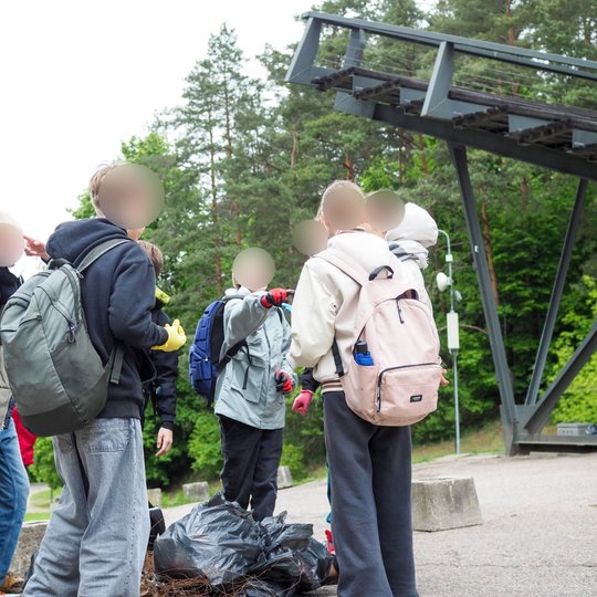 Vaikai stovi ratu šalia sukrautų šiukšlių maišų prie lauko scenos konstrukcijos. Jie dėvi kuprines ir kalbasi po atlikto darbo gamtoje.