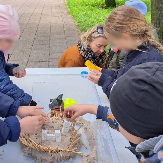 Vaikai susibūrę aplink plastikinę dėžę, kurioje kuria smėlio ir kitų medžiagų pilies maketą su bokštais, tvorele ir vėliava. Veiksmas vyksta lauke, šalia tako ir žalios vejos.