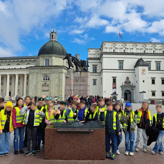 rečioji mokinių grupė taip pat pozuoja prie paminklo Gediminui, šalia taktilinio Vilniaus pilių maketo. Vaikai vilki ryškias liemenes, šypsosi, mojuoja.