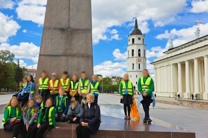 Nuotraukoje – vaikų grupė su lydinčia mokytoja, pozuojanti prie paminklo Vilniaus Katedros aikštėje. Mokiniai dėvi ryškias saugos liemenes, o už jų stūkso istoriniai sostinės simboliai – Katedros varpinė ir pati Vilniaus arkikatedra bazilika su baltomis k