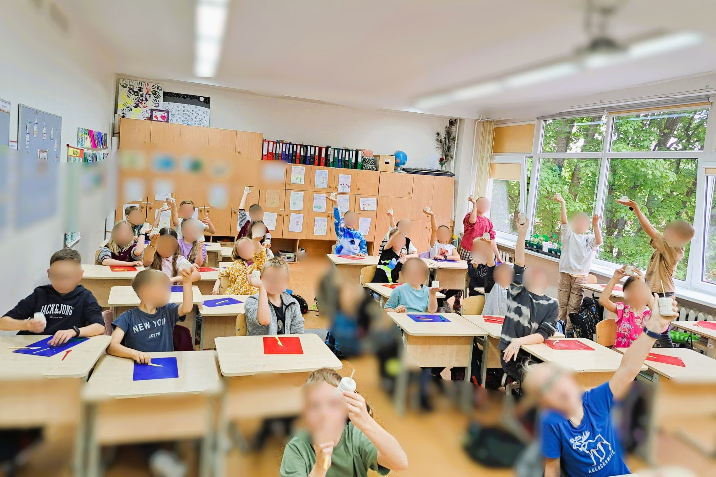 Klasė su iškeltais darbais. Grupė vaikų išdidžiai demonstruoja savo kūrybinius bokštus. Ant sienų – vaikų piešiniai, darbai, segtuvai, o už lango – žaluma. Atmosfera – pilna gyvybės, smalsumo ir pasididžiavimo savo pasiekimais. Kiekvienas 