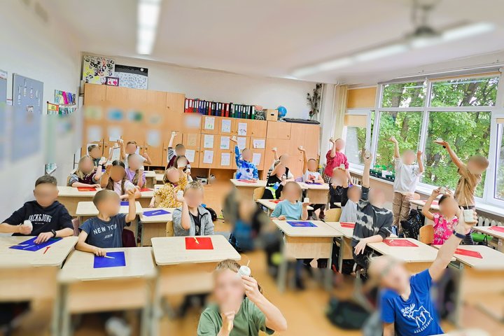 Klasė su iškeltais darbais. Grupė vaikų išdidžiai demonstruoja savo kūrybinius bokštus. Ant sienų – vaikų piešiniai, darbai, segtuvai, o už lango – žaluma. Atmosfera – pilna gyvybės, smalsumo ir pasididžiavimo savo pasiekimais. Kiekvienas