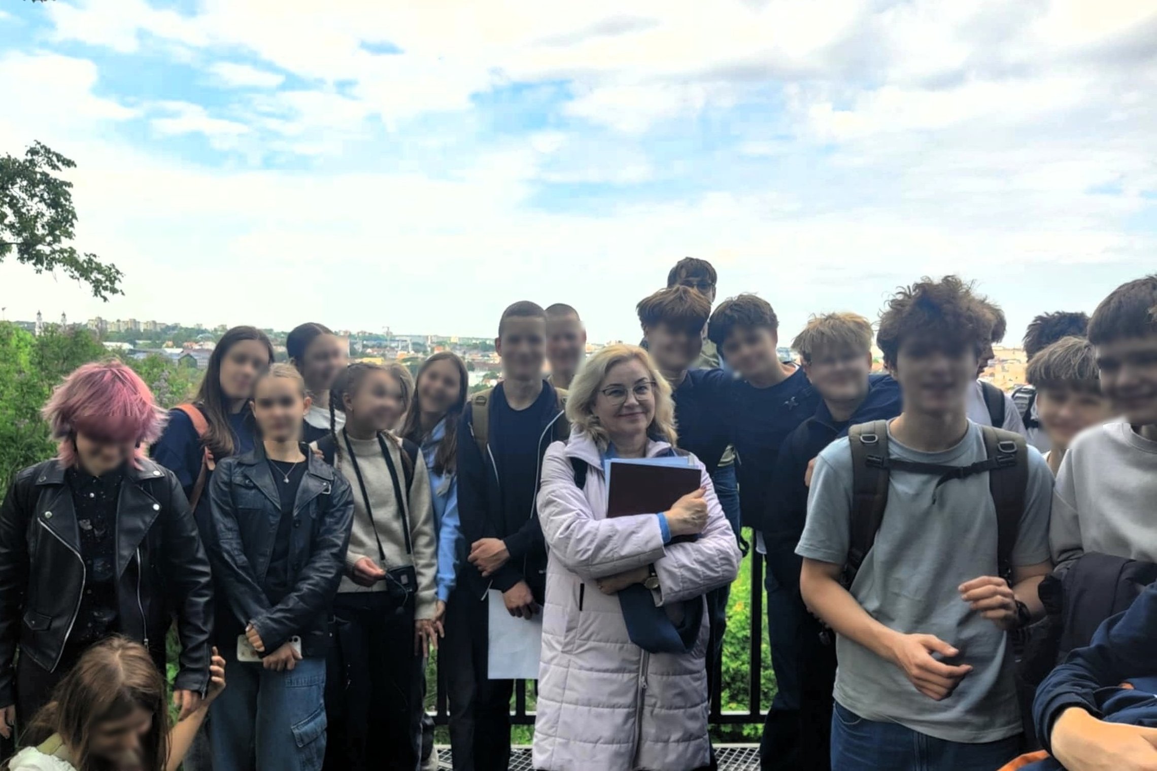 Nuotraukoje matyti grupė paauglių ir viena mokytoja. Jie stovi lauke, ant apžvalgos aikštelės, už jų atsiveria miesto panorama. Dangus debesuotas, bet šviesus – atrodo, kad oras yra palankus pasivaikščiojimui.