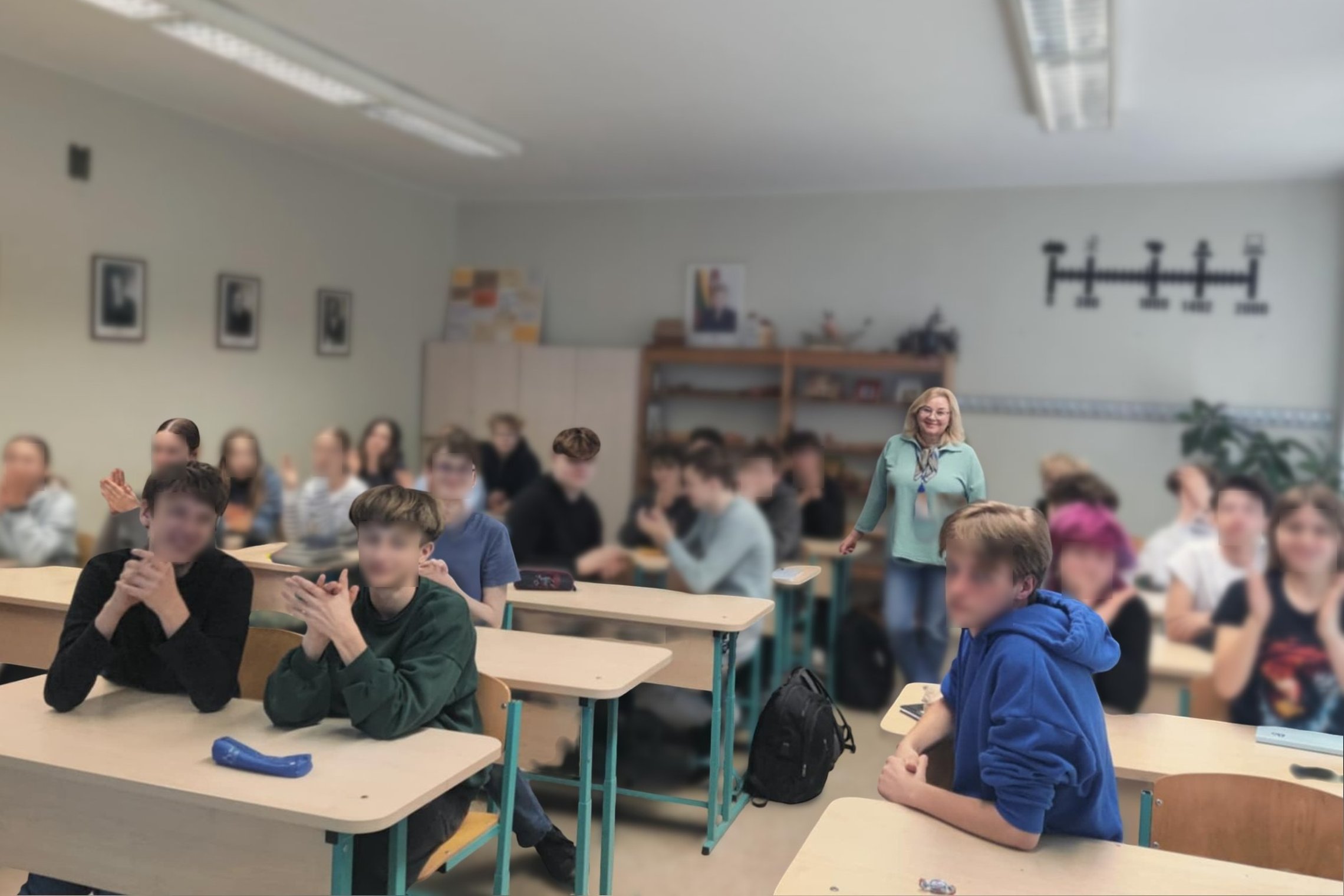 Mokytoja stovi klasės viduryje, o priešais ją – kelios eilės mokinių, sėdinčių prie suolų. Dauguma mokinių atsisukę į kamerą, kai kurie ploja. Klasės aplinka įprasta: šviesios sienos, spintelės, knygų lentynos, ant sienos kabantys paveikslai ir laiko juos