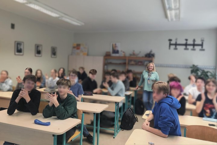 Mokytoja stovi klasės viduryje, o priešais ją – kelios eilės mokinių, sėdinčių prie suolų. Dauguma mokinių atsisukę į kamerą, kai kurie ploja. Klasės aplinka įprasta: šviesios sienos, spintelės, knygų lentynos, ant sienos kabantys paveikslai ir laiko juos