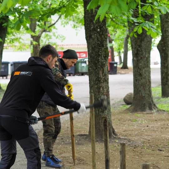 Du vyrai parke montuoja medinę tvorą, vienas jų kala kuolą su kūju. Aplink juos daug medžių, o fone matyti šiukšliadėžės ir pastatai.