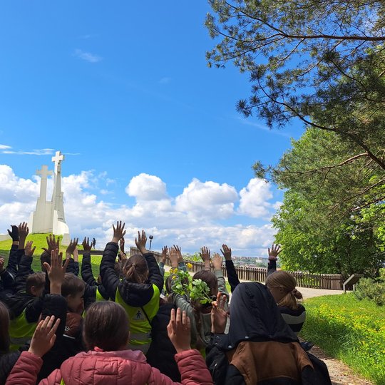 Vaikai su ryškiaspalvėmis liemenėmis pakėlę rankas stovi priešais Trijų Kryžių paminklą, apsupti žalios gamtos ir mėlyno dangaus su debesimis.