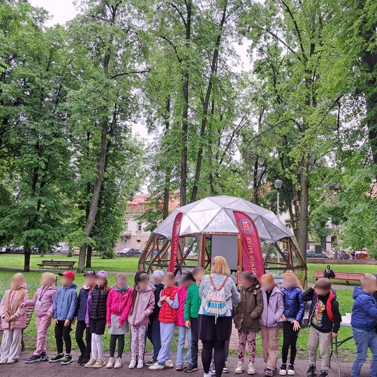 Nuotraukoje – edukacinė veikla vykstanti lauke, Vilniaus pilių teritorijoje, prie pat Gedimino pilies kalno. Vaikai sustoję vienas šalia kito, stovi prie Vilniaus pilių kupolo. Jie ruošiasi statyti piliakalnių modelius nedidelėse grupėse. 
