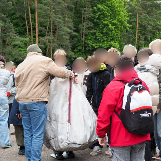 Vaikai ir suaugęs asmuo kartu neša didelį baltą maišą Kalnų parke. Jie stovi grupėje po šiukšlių rinkimo akcijos, fone matyti žali medžiai ir lauko tribūnos.