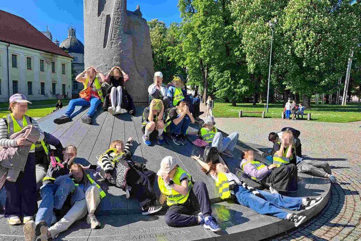 **Alt tekstas:** Trečiokų grupė su ryškiaspalvėmis liemenėmis ilsisi ant paminklo Gediminui prie Vilniaus katedros aikštės, fone matyti istorinis pastatas ir žaliuojantys medžiai.