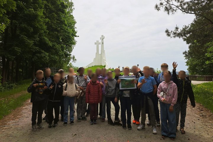 Nuotraukoje – vaikų grupė, pozuojanti Trijų kryžių kalno fone Vilniuje. Tai viena iš žymiausių sostinės vietų, dažnai lankoma ekskursijų metu. Vaikai stovi miško apsuptyje, kai kurie laiko piešinį ar plakatą – tikėtina, tai pažintinės ar edukacinės išvyko