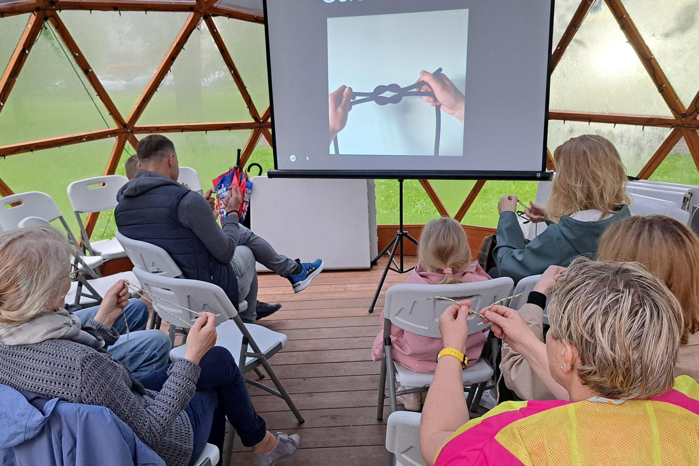 Dalyviai kupolo viduje seka instrukcijas iš ekrano ir mokosi rišti mazgus, laikydami virves rankose. Už langų matyti žalias parkas ir lietaus palikti lašai ant kupolo sienelių.