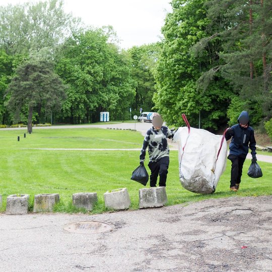 Du vaikai tempia didelį baltą šiukšlių maišą ir neša juodus maišelius per pievą. Fone matyti žalios vejos, takeliai ir parką supantys medžiai.