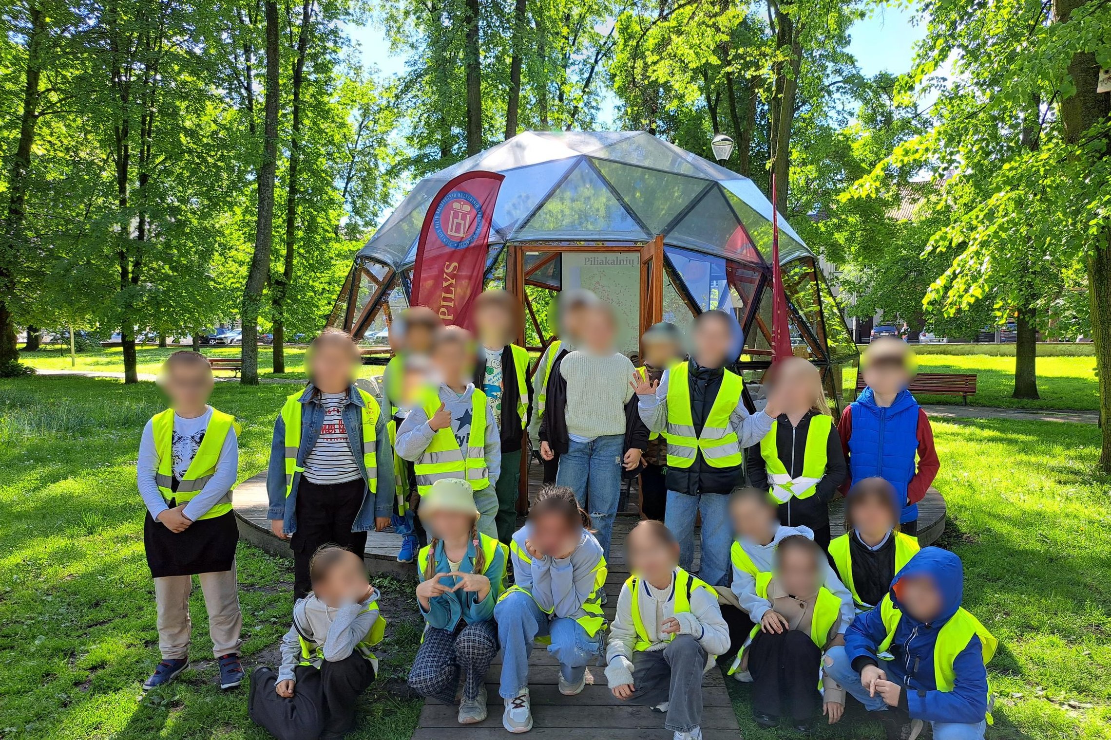 Nuotraukoje – grupė vaikų, pozuojanti prie kupolo formos stiklinės pavėsinės žalioje, medžiais apsuptoje aplinkoje. Visi vaikai dėvi ryškias geltonas saugos liemenes, o už jų matomas stendas su užrašu „Piliakalniai“ bei raudonas stovas su vėliava „VILNIAU
