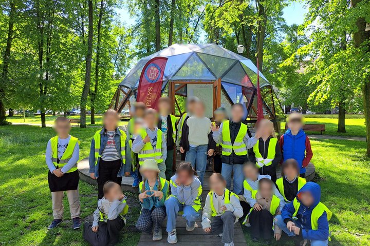 Nuotraukoje – grupė vaikų, pozuojanti prie kupolo formos stiklinės pavėsinės žalioje, medžiais apsuptoje aplinkoje. Visi vaikai dėvi ryškias geltonas saugos liemenes, o už jų matomas stendas su užrašu „Piliakalniai“ bei raudonas stovas su vėliava „VILNIAU