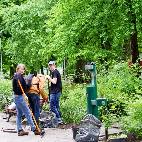 Trys žmonės valo krūmynus parke, šalia jų stovi šiukšlių maišai ir įrankiai. Priekyje matoma vandens gėrimo stotelė, fone – medžiai ir raudona palapinė.