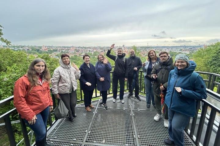 Grupė devynių žmonių stovi Altanos bokšto terasoje, fone matyti Vilniaus senamiesčio panorama su raudonais stogais, žaliais medžiais ir Televizijos bokštu tolumoje. Dalis žmonių šypsosi, mojuoja ar laiko skėčius, visi apsirengę šiltai – matyti striukės,
