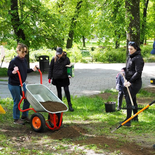 Keturi žmonės parke tvarko žemę: viena moteris pila žemes iš karučio, o kitos grėbia ir sėja. Veiksmas vyksta šalia tako, apsupto žaliuojančių medžių.