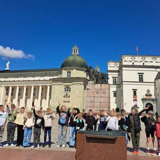 Kita didelė mokinių grupė stovi Katedros aikštėje prie paminklo Gediminui. Už jų – Vilniaus arkikatedra bazilika ir Valdovų rūmai. Vaikai kelia rankas, šypsosi – akivaizdu, kad nuotaika puiki