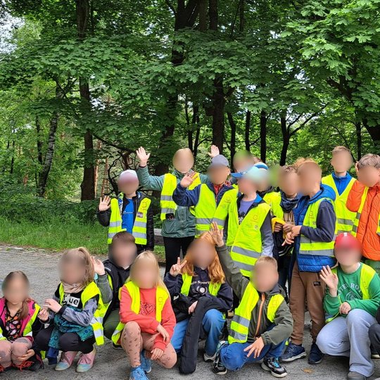 nuotraukoje – sustojusi vaikų grupė po medžių lajomis. Jie šypsosi, mojuoja, pozuoja nuotraukai kartu su lydinčiu suaugusiuoju. Fone – tankus miškas, sukuriantis jaukią ir draugišką atmosferą.
