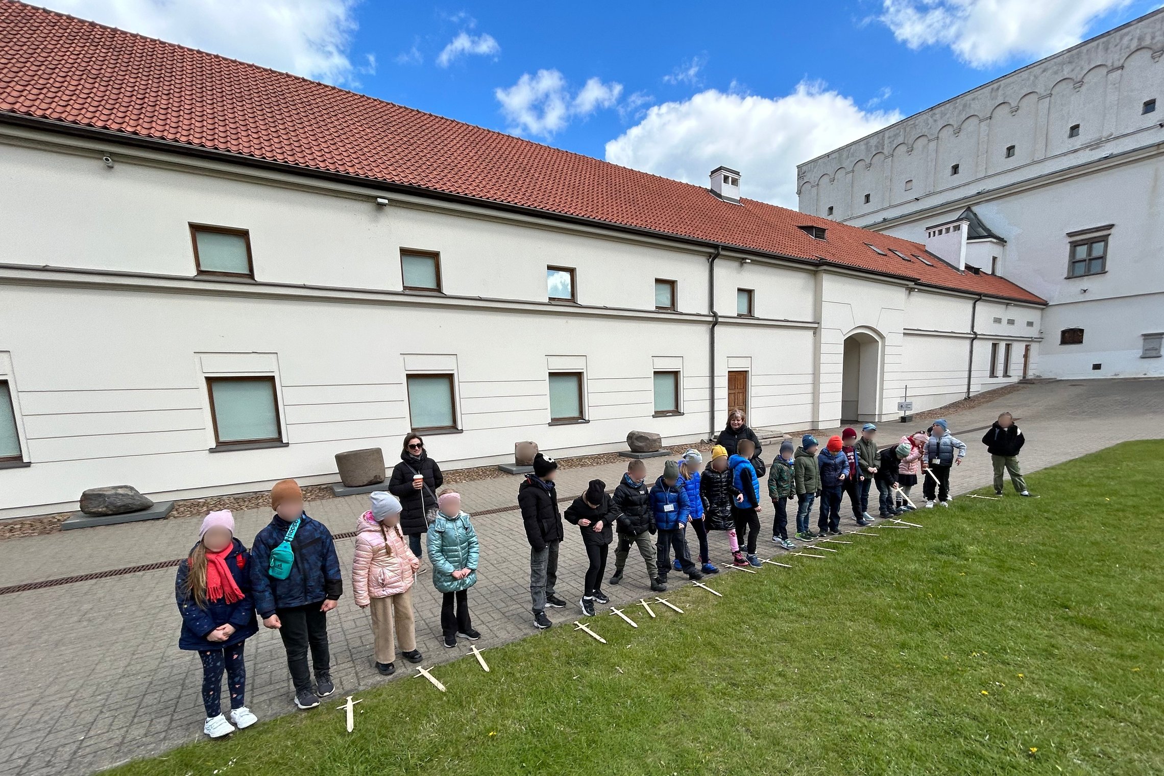 Grupė vaikų stovi eilėje lauke prie arsenalo pastato su raudonu čerpių stogu ir baltomis sienomis. Prie kiekvieno vaiko kojų padėta po medinį kalaviją. Už vaikų stovi dvi mokytojos. Dangus dalinai debesuotas, oras vėsus – vaikai dėvi kepures ir striukes.