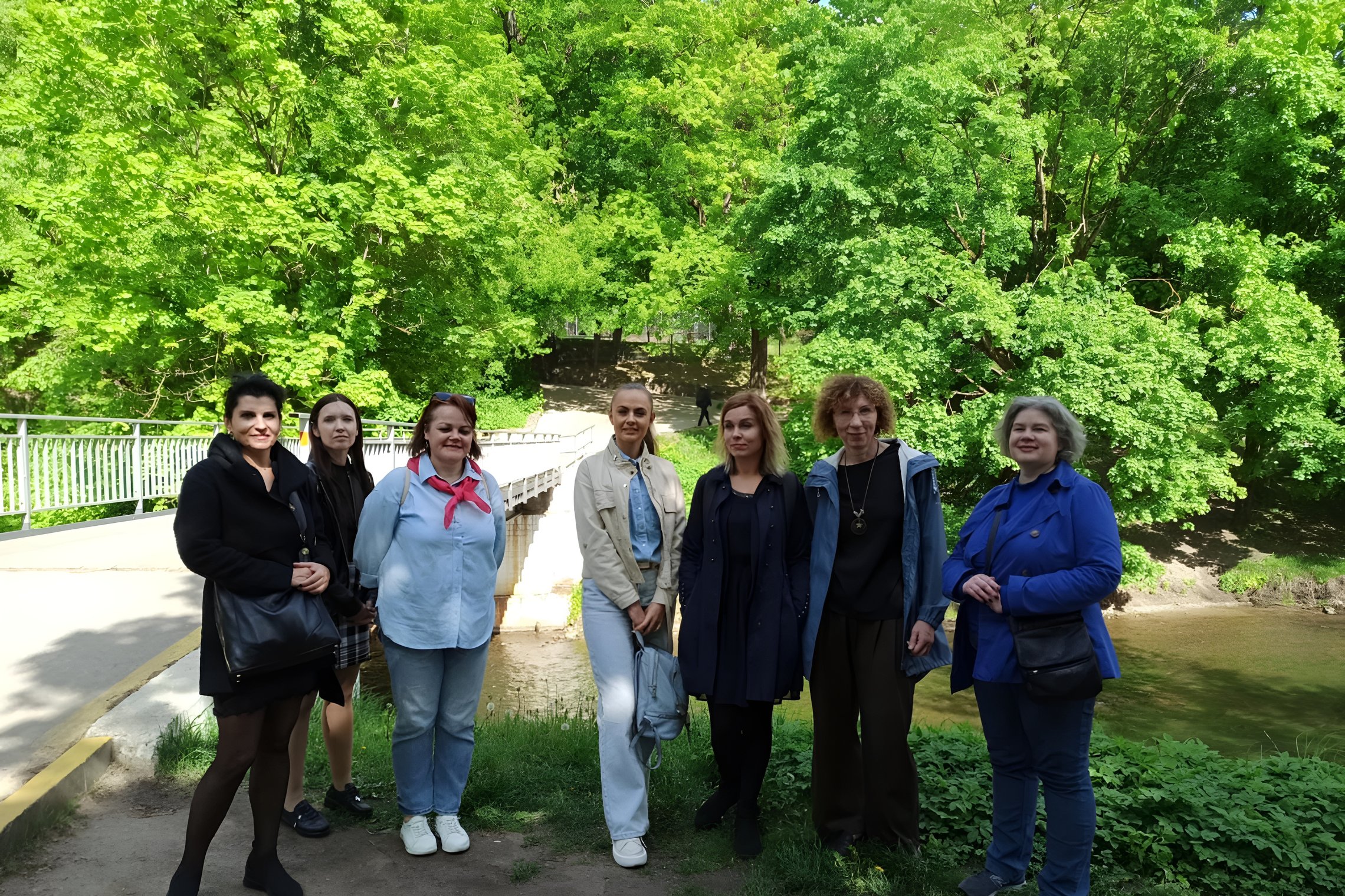 Grupė moterų stovi gamtos apsuptyje prie pėsčiųjų tilto per upelį. Fone – sodriai žali medžiai, šviesi pavasario diena. Moterų apranga rodo, kad tai kolektyvinė ekskursija. Kai kurios šypsosi, kitos žvelgia į objektyvą rimtai.