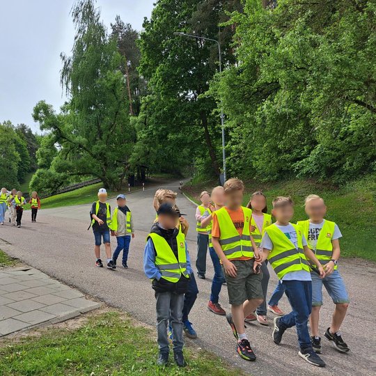 nuotraukoje – vaikai žygiuoja parko taku, pasklidę į mažesnes grupeles. Kai kurie žvalgosi, kalbasi, kiti noriai žingsniuoja pirmyn. Ši scena parodo ekskursijos dinamiką, judėjimą tarp veiklų.