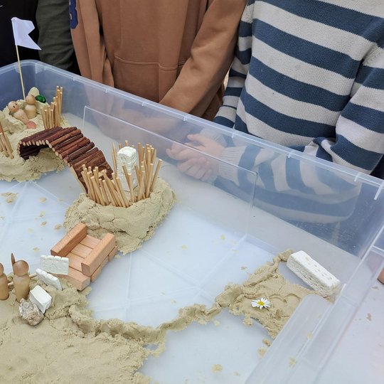 Nuotraukoje – vienas iš vaikų sukurtų piliakalnių modelių, pastatytas edukacinės veiklos metu. Jis sumaniai suformuotas iš smėlio ir įvairių medžiagų: medinių pagaliukų, kaladėlių, figūrėlių bei natūralių elementų, tokių kaip akmenukai ir gėlės.
