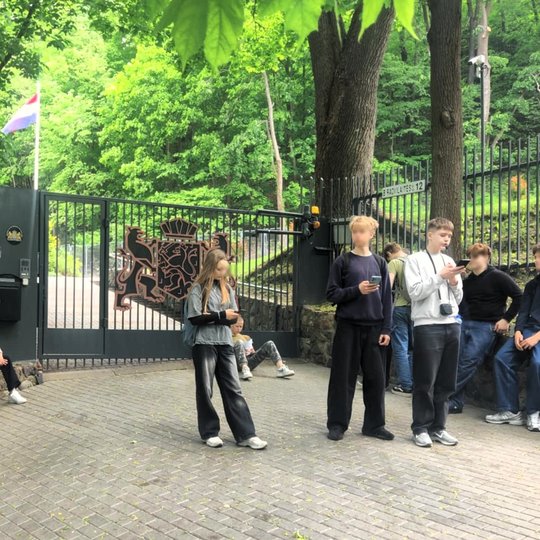 Grupė paauglių mokinių ilsisi ir bendrauja prie Nyderlandų Karalystės ambasados Vilniuje, adresu B. Radvilaitės gatvė 12. Dešinėje kadro pusėje ant metalinės tvoros matyti adreso lentelė, o už jų stovi masyvūs pilki vartai, papuošti iškiliu Nyderlandų kar
