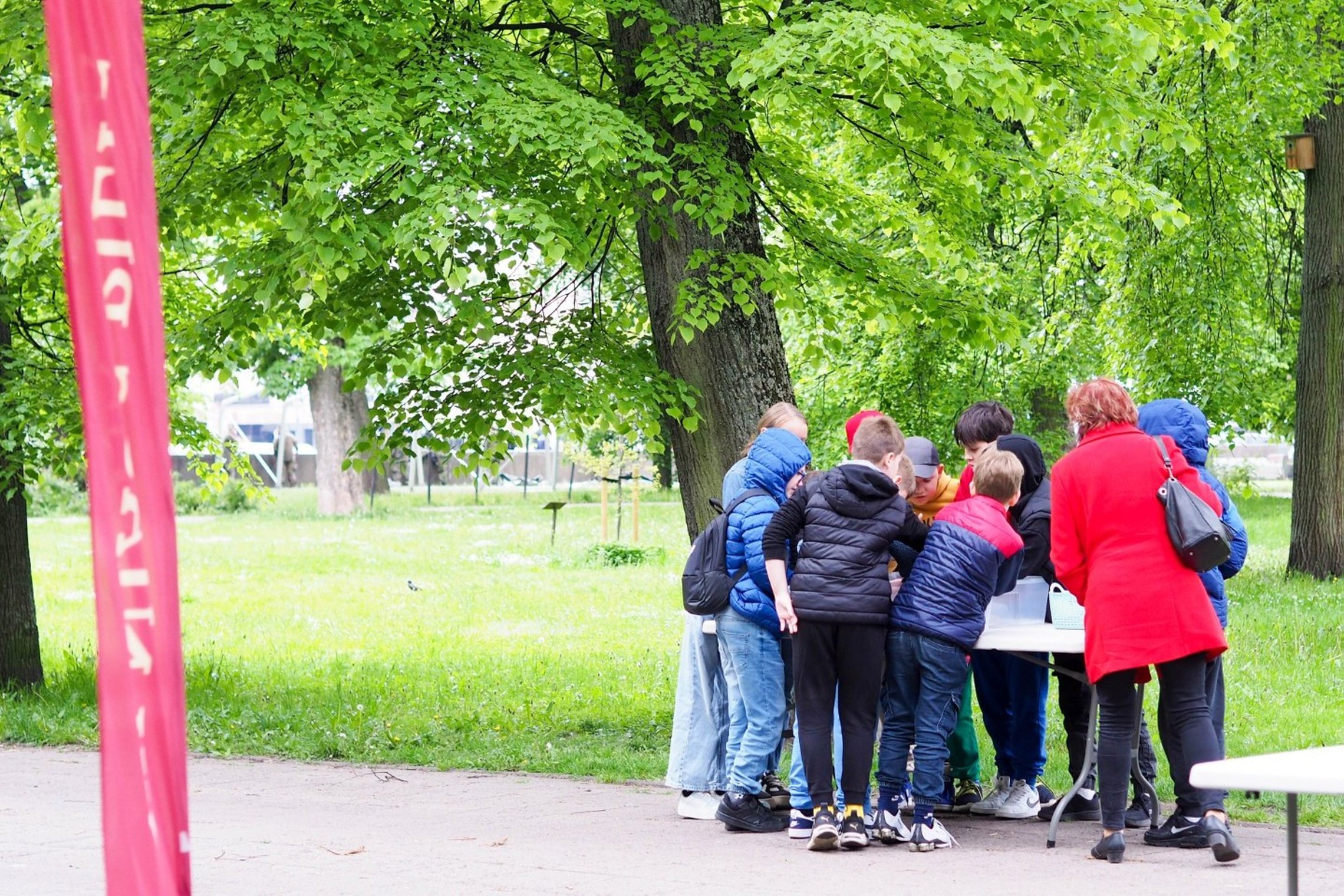 Vaikai ir mokytoja lauke prie stalo susibūrę edukacinėje veikloje, apsirengę šilčiau, žalių medžių fone, dalyvauja edukacijoje.