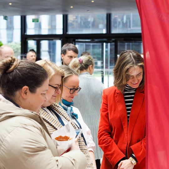 Grupė moterų stovi prie informacinio stendo su raudona vėliava, ant kurios matomas užrašas „Vilniaus pilys“. Jos įdėmiai žiūri į kažką stende. Tarp jų – moteris su ryškiu raudonu švarku ir dryžuotu golfu, besišypsanti ir palinkusi į priekį. Fone matyti da