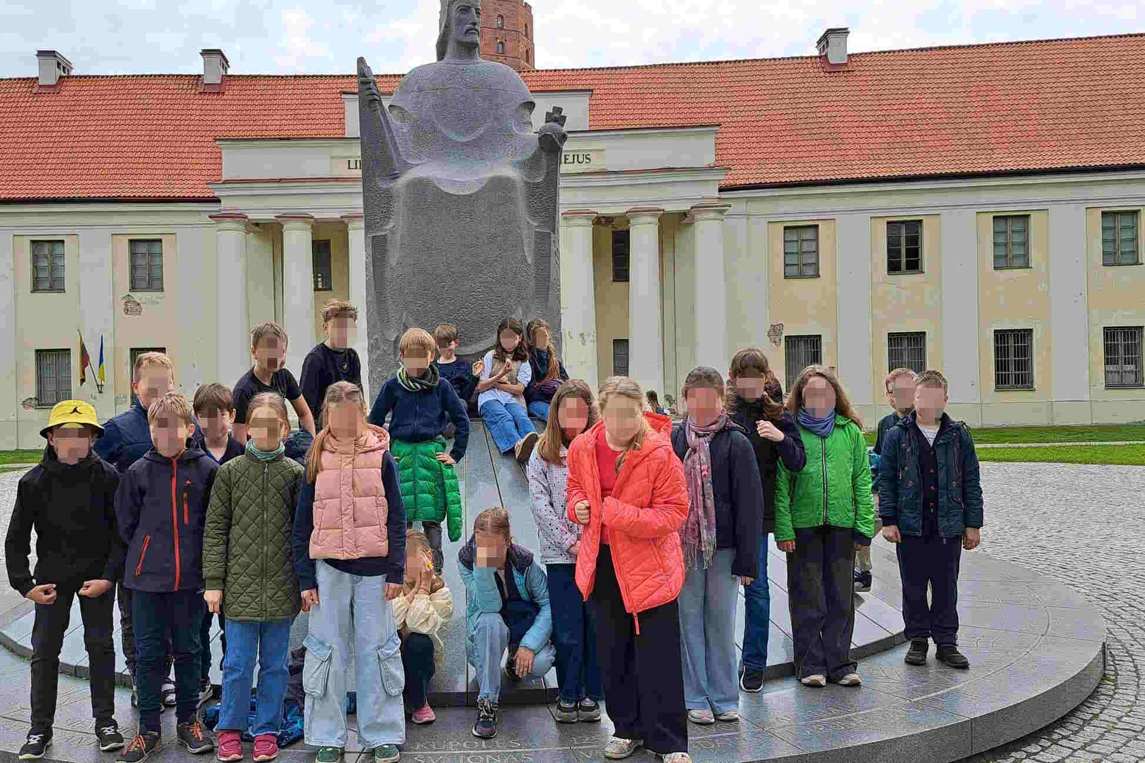 Grupė vaikų pozuoja prie Lietuvos didžiojo kunigaikščio Gedimino paminklo Vilniaus pilių teritorijoje, fone – Senieji arsenalai su raudonu stogu ir užrašu „Lietuvos nacionalinis muziejus“.
