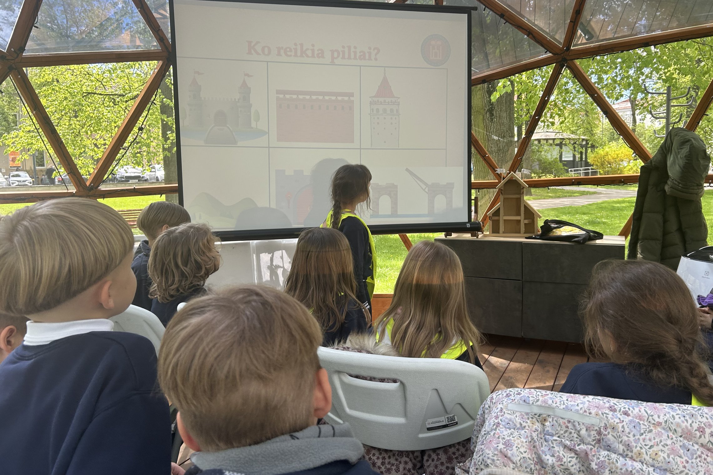 Grupė pradinukų dalyvauja edukacijoje kupole. Vaikai sėdi plastikinėse kėdėse ir žiūri į projektoriaus ekraną, kuriame rodomas skaidrių pristatymas pavadinimu „Ko reikia piliai?“ ir pilies elementų iliustracijomis. Viena mergaitė stovi prie ekrano. Fone –