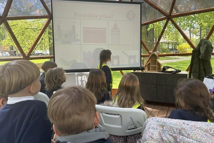 Grupė pradinukų dalyvauja edukacijoje kupole. Vaikai sėdi plastikinėse kėdėse ir žiūri į projektoriaus ekraną, kuriame rodomas skaidrių pristatymas pavadinimu „Ko reikia piliai?“ ir pilies elementų iliustracijomis. Viena mergaitė stovi prie ekrano. Fone –