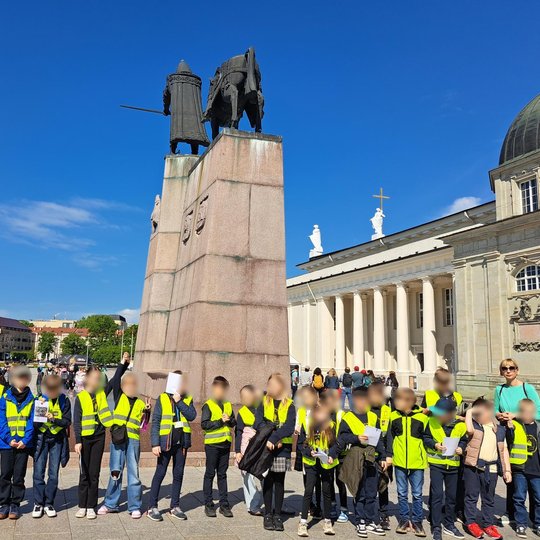 Mokinių grupė su šviesą atspindinčiomis liemenėmis stovi šalia paminklo Gediminui, kai kurie laiko užduočių lapus. Fone – Katedros aikštė, paminklas, ir Vilniaus arkikatedra.