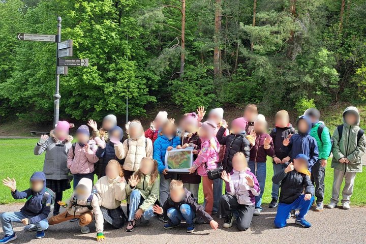 Vaikų grupė pozuoja prie kelio ženklų su nuorodomis į Vilniaus istorines vietas; už jų – žaliuojantis miškas. Vaikai šypsosi ir mojuoja, laikydami ekskursijos žemėlapį.