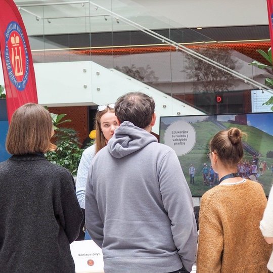 Grupė žmonių stovi prie informacinio stendo, pažymėto raudonomis vėliavomis su Vilniaus pilių logotipu. Centre – ekranas, kuriame matomas Gedimino kalnas ir užrašas „Edukacijos su vaizdu į valstybės pradžią“. Viena iš stendo atstovių kalbasi su lankytojai