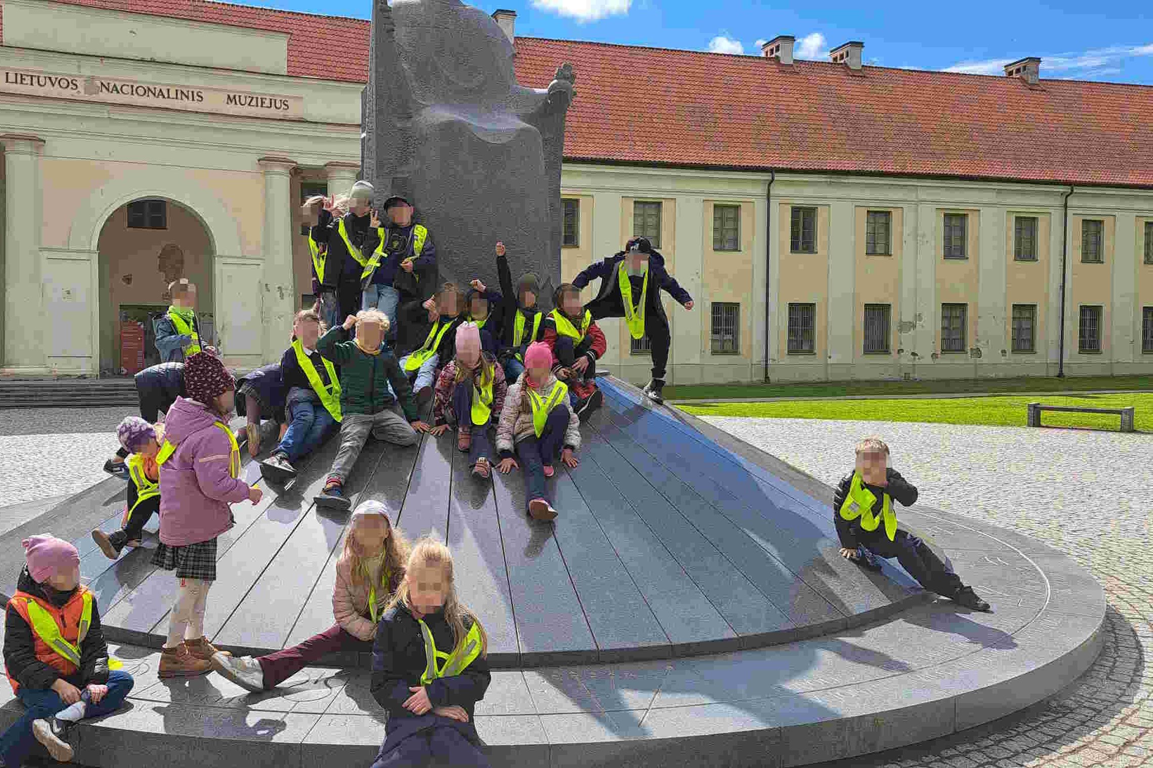 Vaikai su ryškiaspalvėmis liemenėmis žaidžia prie Karaliaus Mindaugo paminklo Vilniuje, šalia Lietuvos nacionalinio muziejaus. Saulėtą dieną grupė atsipalaidavusi po edukacinės veiklos.